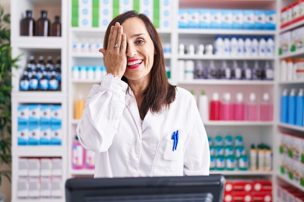 Foto gratuita mujer morena de mediana edad que trabaja en la farmacia que cubre un ojo con una sonrisa segura en la cara y emoción sorpresa