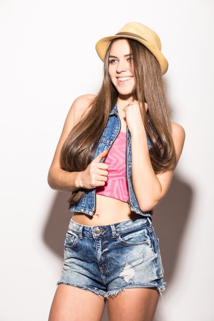 Mujer morena de moda con sombrero de paja y gafas de sol sonriendo aislado sobre pared blanca