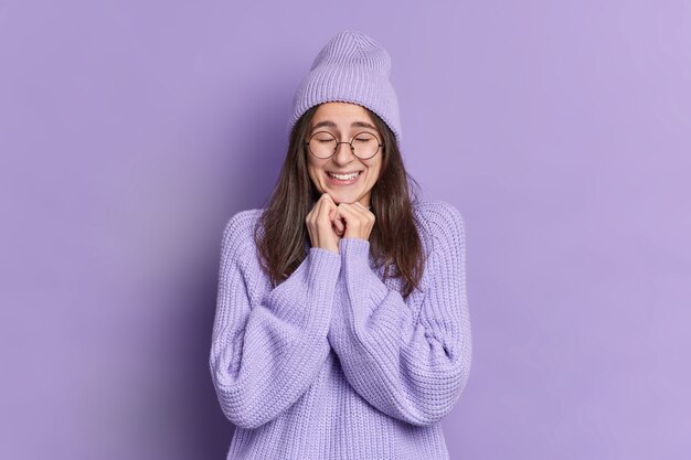 Mujer morena muy complacida mantiene las manos debajo de la barbilla, cierra los ojos y se siente encantada de escuchar algo bueno vestida con anteojos grandes de sombrero de puente de invierno cálido.