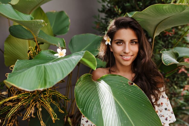 Mujer morena de ojos verdes con flor en el pelo mira al frente, posando entre grandes hojas de plantas tropicales