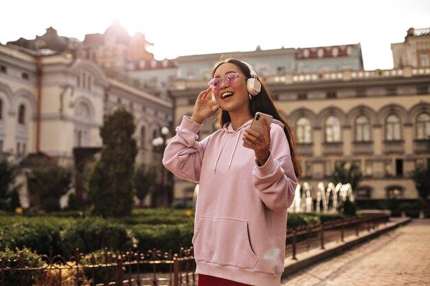 Mujer morena optimista con elegante sudadera con capucha y gafas de sol rosas cantando, sosteniendo el teléfono y escuchando música en auriculares afuera