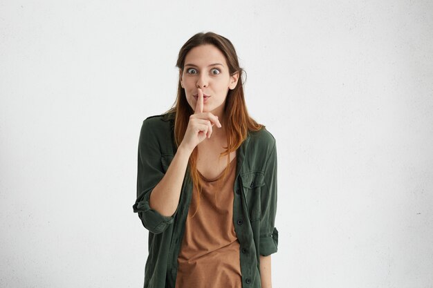 Mujer morena con pelo lacio con chaqueta verde manteniendo su dedo índice en los labios haciendo señal de silencio pidiendo no ser ruidosa.