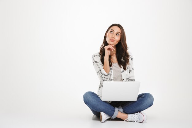 Mujer morena pensativa en camisa sentada en el suelo con la computadora portátil y mirando hacia arriba sobre gris