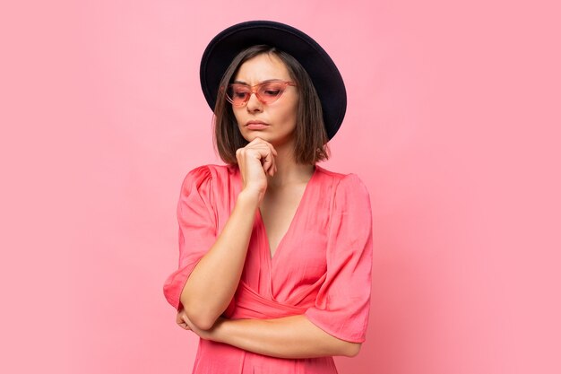 Mujer morena pensativa en elegantes gafas posando sobre pared rosa.