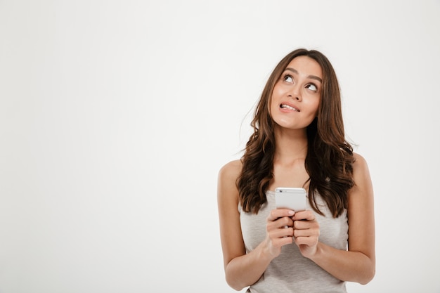 Mujer morena pensativa que sostiene el teléfono inteligente y mirando hacia arriba sobre gris