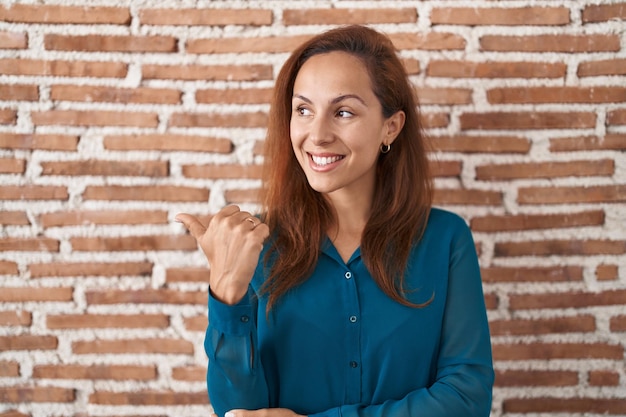 Mujer morena de pie sobre la pared de ladrillos sonriendo con cara feliz mirando y apuntando hacia un lado con el pulgar hacia arriba