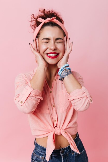 Mujer morena positiva en blusa rosa y diadema estilo pinup sonríe con los ojos cerrados y toca su cara sobre fondo rosa