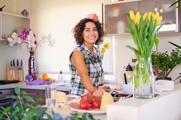 Mujer morena positiva con cabello rizado hace ensalada con tomates y papas en una cocina casera.