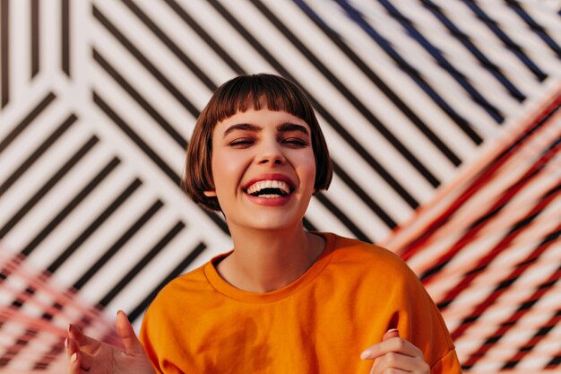 Mujer morena positiva riendo sinceramente sobre fondo rayado Chica de pelo corto con sudadera naranja sonriendo al aire libre