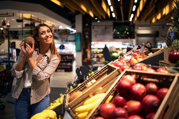 Mujer morena positiva sosteniendo coco en el departamento de frutas de la tienda de comestibles