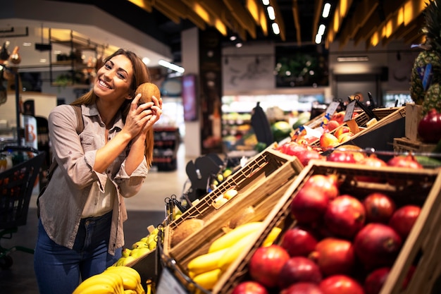 Mujer morena positiva sosteniendo coco en el departamento de frutas de la tienda de comestibles