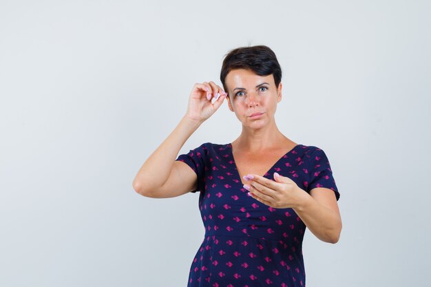 Mujer morena que finge depilarse las cejas, mirar al espejo con un vestido estampado morado y rojo y mirar concentrada. vista frontal.