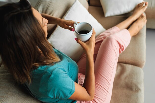 Mujer morena en ropa de hogar acogedor con taza de té, mirando a la ventana y relajarse en el sofá moderno