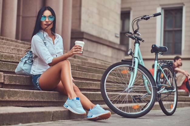 Una mujer morena sexy con blusa y pantalones cortos de mezclilla con gafas de sol, relajándose después de andar en bicicleta, sentada con una taza de café en los escalones de una ciudad.