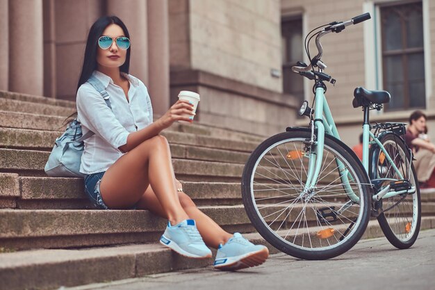 Una mujer morena sexy con blusa y pantalones cortos de mezclilla con gafas de sol, relajándose después de andar en bicicleta, sentada con una taza de café en los escalones de una ciudad.