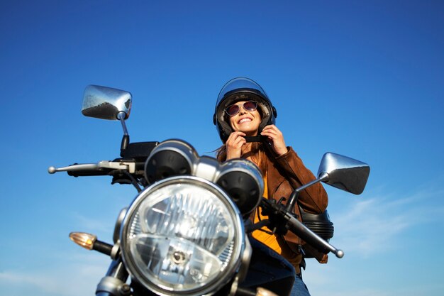 Mujer morena sexy en chaqueta de cuero poniéndose el casco y sentado en una motocicleta de estilo retro en un hermoso día soleado