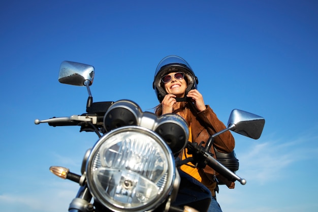 Mujer morena sexy en chaqueta de cuero poniéndose el casco y sentado en una motocicleta de estilo retro en un hermoso día soleado