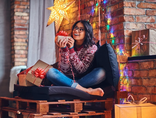 Mujer morena sexy con los pies desnudos vestida con jeans y un suéter rojo posando en un sofá en una habitación con decoración navideña.