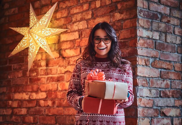 Mujer morena sonriente con anteojos y un suéter cálido sostiene regalos de Navidad sobre la pared de un ladrillo.