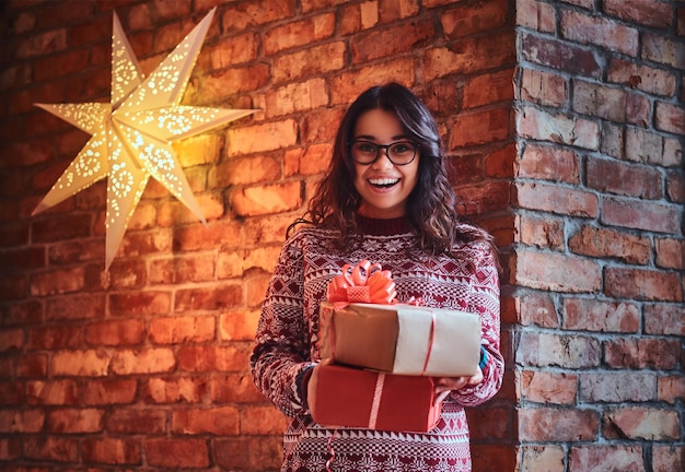 Mujer morena sonriente con anteojos y un suéter cálido sostiene regalos de Navidad sobre la pared de un ladrillo.