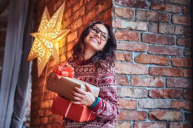 Mujer morena sonriente con anteojos y un suéter cálido sostiene regalos de Navidad sobre la pared de un ladrillo.