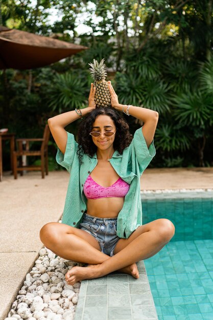 Mujer morena sonriente posando con piña sentada cerca de la piscina en un resort tropical
