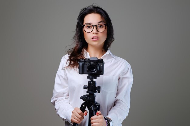 Mujer morena sonriente y positiva con anteojos tomando fotos con una cámara fotográfica profesional. Aislado sobre fondo gris.