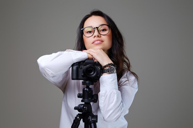 Mujer morena sonriente y positiva con anteojos tomando fotos con una cámara fotográfica profesional. Aislado sobre fondo gris.