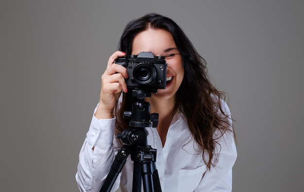 Mujer morena sonriente y positiva con anteojos tomando fotos con una cámara fotográfica profesional. Aislado sobre fondo gris.