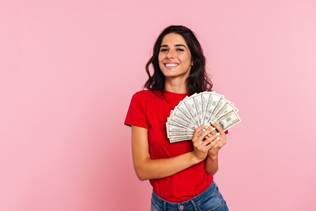 Mujer morena sonriente que sostiene el dinero en manos y que mira la cámara sobre rosa