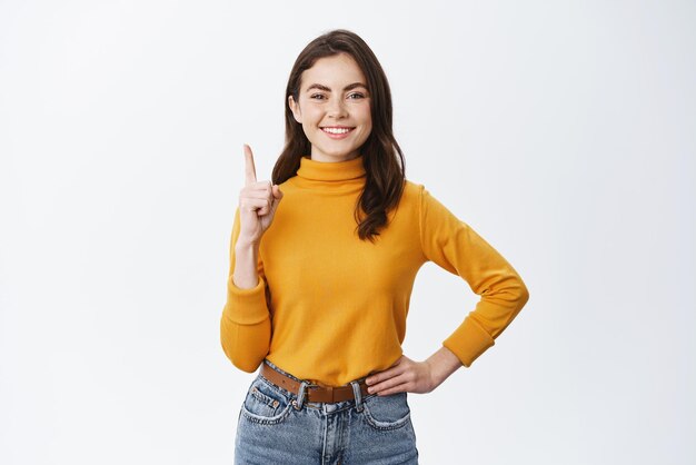 Mujer morena sonriente en suéter amarillo apuntando con el dedo hacia arriba Modelo femenino feliz que muestra un anuncio en la parte superior que indica en el espacio de la copia de pie sobre fondo blanco