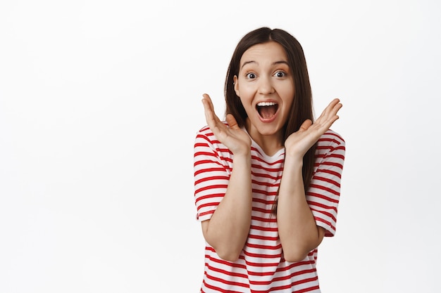 Mujer morena sorprendida y emocionada, grito de felicidad, ganando, triunfando y celebrando, de pie en camiseta contra la pared blanca