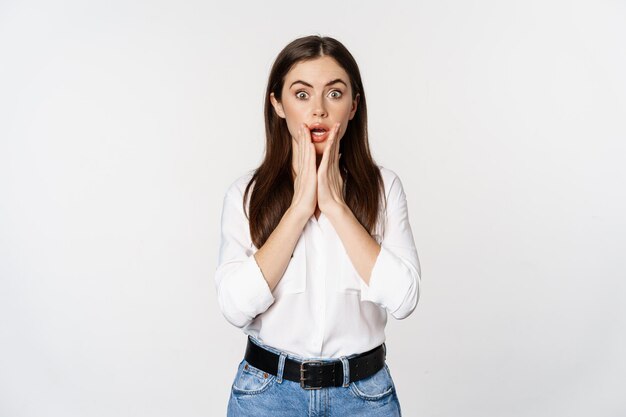 Mujer morena sorprendida mirando asombrada, viendo algo increíble, de pie en blusa y jeans sobre fondo blanco.