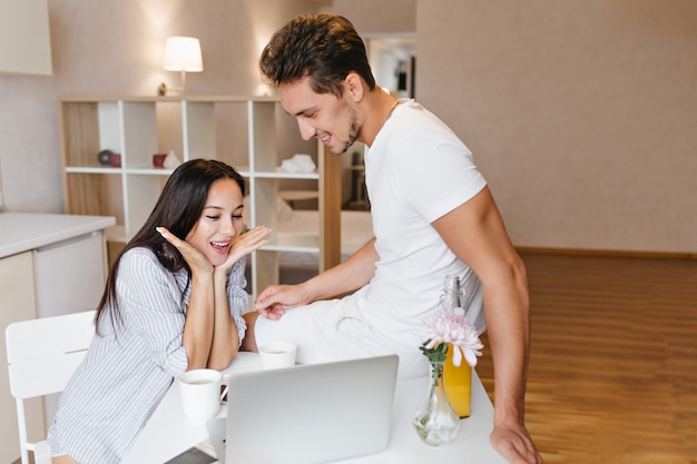 Mujer morena sorprendida sentada cerca de la computadora portátil durante el desayuno con su novio en camiseta blanca