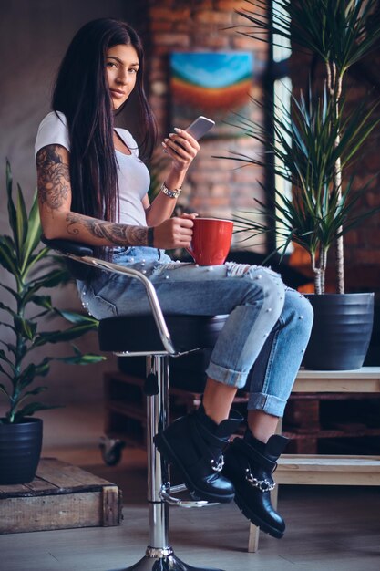 Mujer morena tatuada bebe café caliente en una habitación con interior de loft.