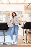 Foto gratuita mujer morena con teléfono parado en cocina panorámica en ropa casual