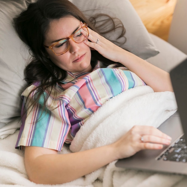 Mujer morena de tiro medio quedándose en la cama y trabajando en la computadora portátil