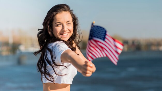 Mujer morena de tiro medio sosteniendo 2 banderas de Estados Unidos cerca del lago