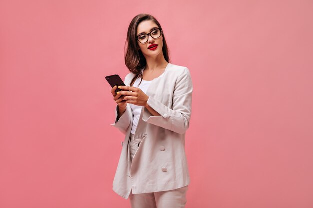 Mujer morena en traje elegante y anteojos posa con smartphone. Mujer de negocios joven con labios rojos y traje blanco sostiene teléfono.
