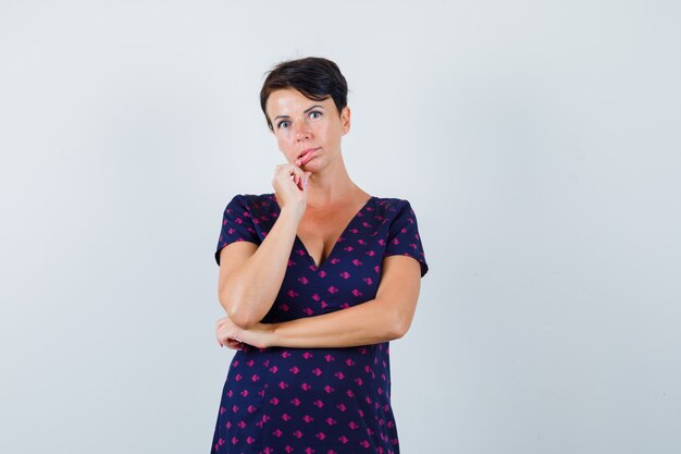 Mujer morena en vestido estampado morado y rojo sosteniendo sus dedos cerca de su boca y pensando en algo y mirando pensativa, vista frontal.