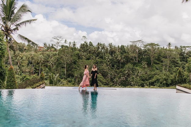 Mujer morena en vestido rosa cogidos de la mano con un amigo en Bali. Foto al aire libre de modelos femeninos de pie junto a la piscina en la selva.