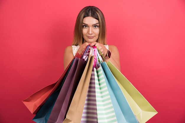 Mujer mostrando bolsas de la compra con expresión neutra en la pared roja.