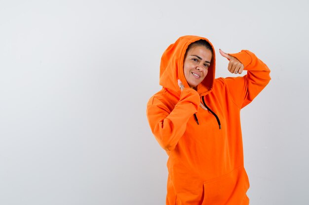 Mujer mostrando doble pulgar hacia arriba en sudadera con capucha naranja y mirando feliz
