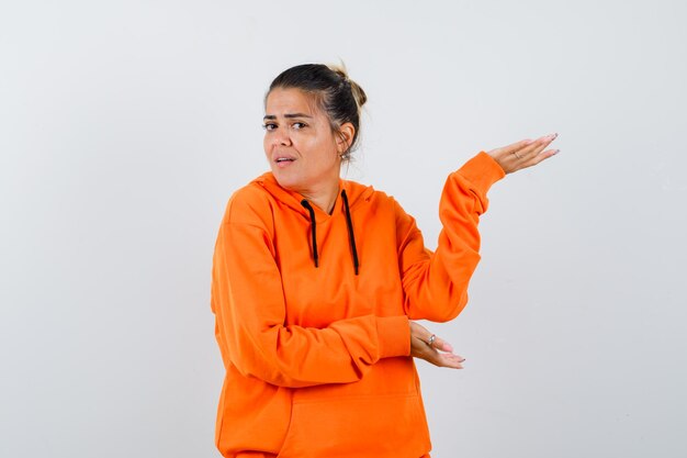 Mujer mostrando gesto de bienvenida en sudadera con capucha naranja y mirando confiado