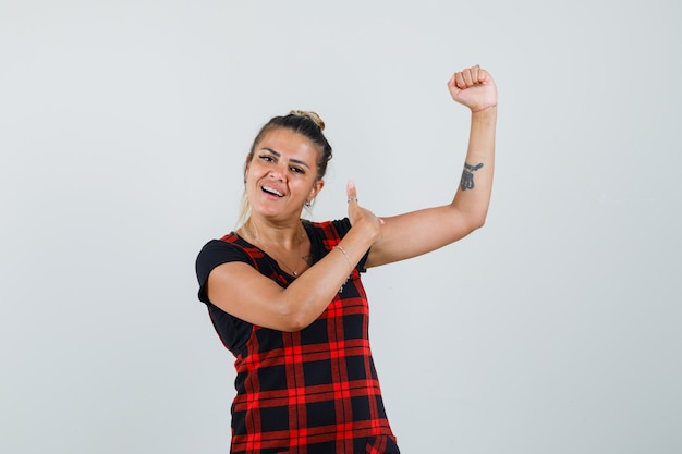 Foto gratuita mujer mostrando los músculos con el pulgar hacia arriba en vestido delantal y luciendo confiada. vista frontal.