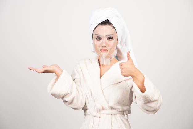 Mujer mostrando el pulgar hacia arriba y vistiendo albornoz y toalla con mascarilla.