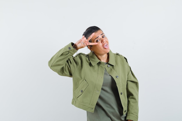 Mujer mostrando el signo V cerca del ojo en chaqueta, camiseta y luciendo confiada