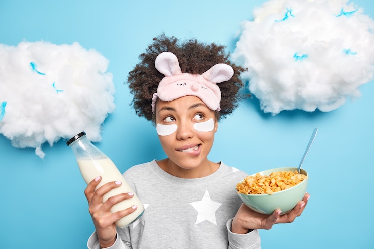 Foto gratuito mujer muerde los labios yendo a desayunar sostiene un tazón de cereales y una botella de leche usa un pijama se aplica parches debajo de los ojos para eliminar las arrugas aisladas en azul