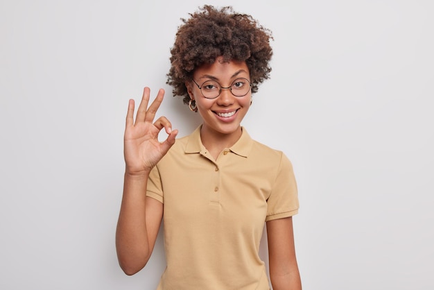 Foto gratuita la mujer muestra un buen gesto con expresión satisfecha aprueba o le gusta algo recomienda algo dice que sí elogia la buena elección usa gafas redondas y una camiseta informal posa en el interior