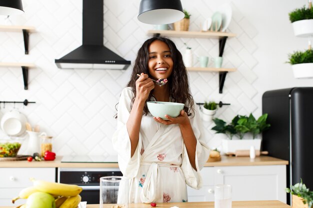 La mujer mulata atractiva sonreída está comiendo frutas cortadas en la cocina moderna blanca vestida con ropa de dormir con el pelo suelto desordenado y mirando directamente